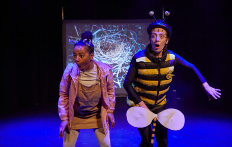 Two performers on stage with their mouths open in surprise, one is dressed as a bee.