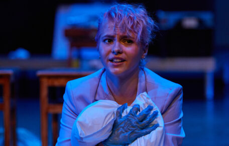 A performer wearing a suit jacket and plastic gloves cradles a sheet in a way reminiscent of holding a baby.