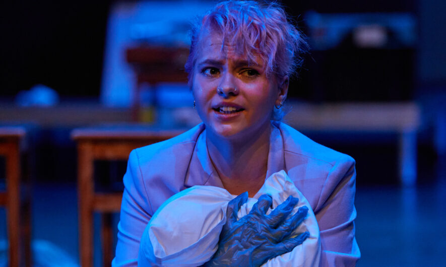 A performer wearing a suit jacket and plastic gloves cradles a sheet in a way reminiscent of holding a baby.
