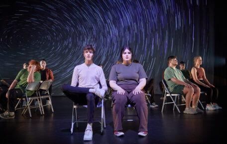 Performers sit on metal chairs on stage facing forward.