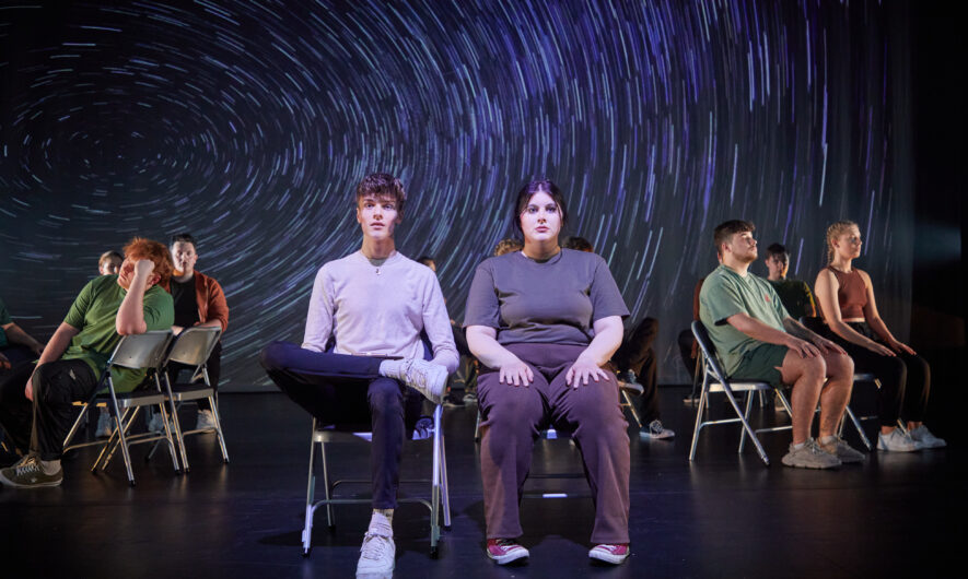 Performers sit on metal chairs on stage facing forward.