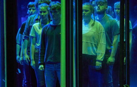 students washed in green and blue light stand behind a glass pane