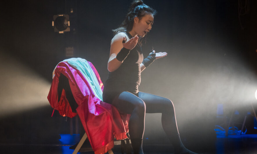 A dancer sits on a chair with a red dress on the back of it. They have their hands up as if to shrug.