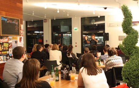 The cafe is filled with people, there's a small stage with a comedian performing.