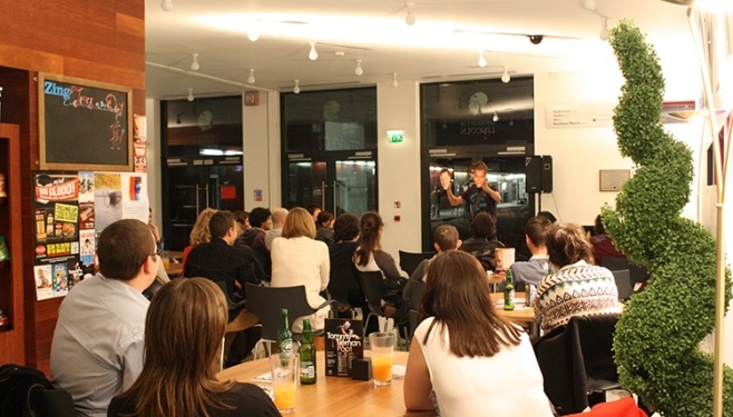 The cafe is filled with people, there's a small stage with a comedian performing.