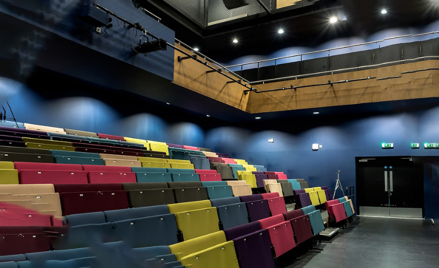 Hire Us, image is of our rainbow coloured double seating. The walls are a deep blue with lights beaming down.