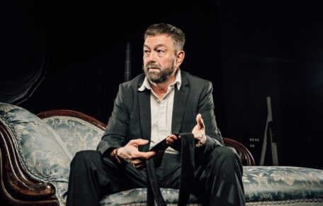 Production image from, My Family and Other Songs. Jed is pictured in a black suit and white shirt and is sat on a sofa holding a tie. Jed has short dark grey hair.