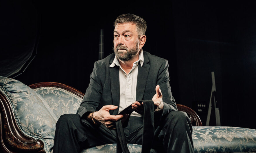Production image from, My Family and Other Songs. Jed is pictured in a black suit and white shirt and is sat on a sofa holding a tie. Jed has short dark grey hair.