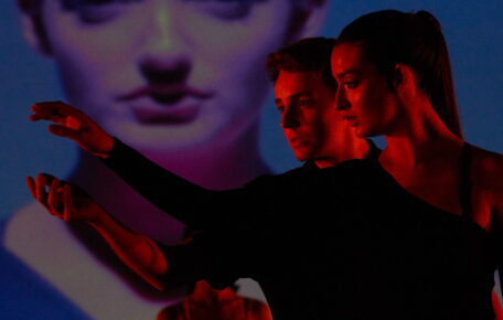 Two people on stage washed in red light both reaching to the left.