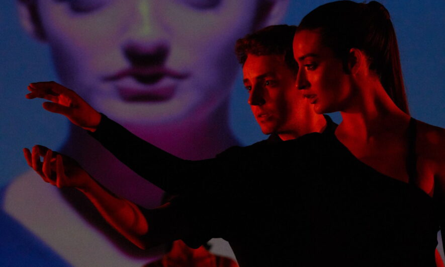 Two people on stage washed in red light both reaching to the left.