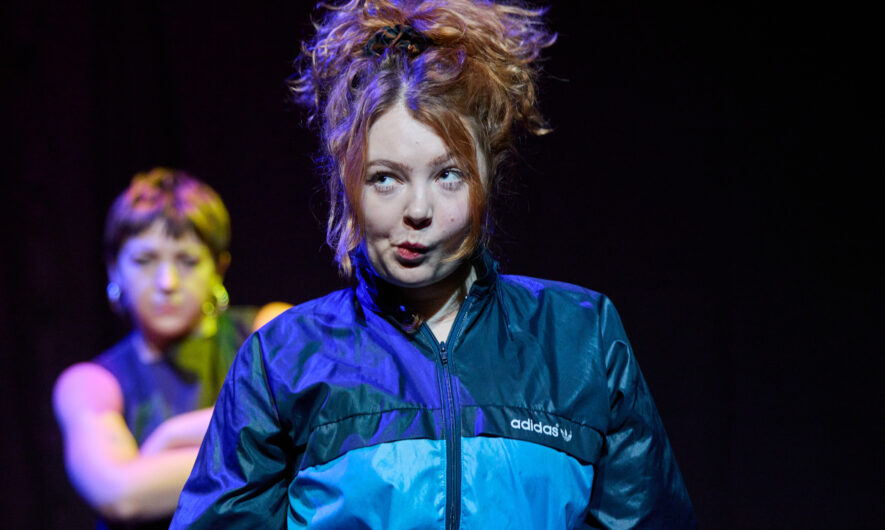 A performer in a blue windbreaker stands centre stage with their lips pursed.