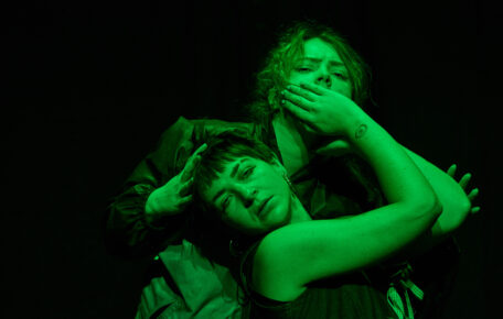 Two performers on stage washed in green light. One holds their hand over the others mouth.