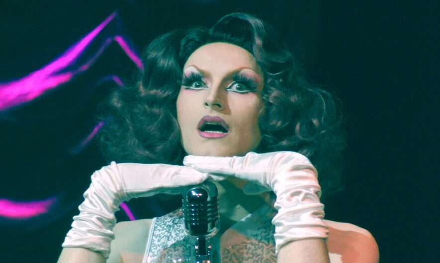 Promotional image for Queen Bayard by Jamie Walsh. A drag performer on stage, with dark hair a white dress and gloves posing over a microphone.