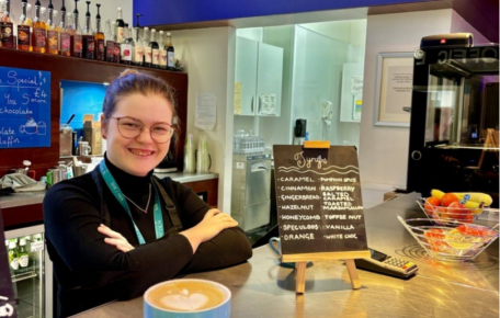 Cafe worker smiling at the camera with a freshly made coffee on the Cafe bar.