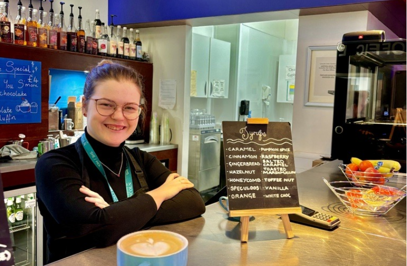 Cafe worker smiling at the camera with a freshly made coffee on the Cafe bar.