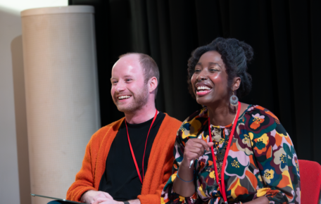 Two people sit and smile towards someone off camera.