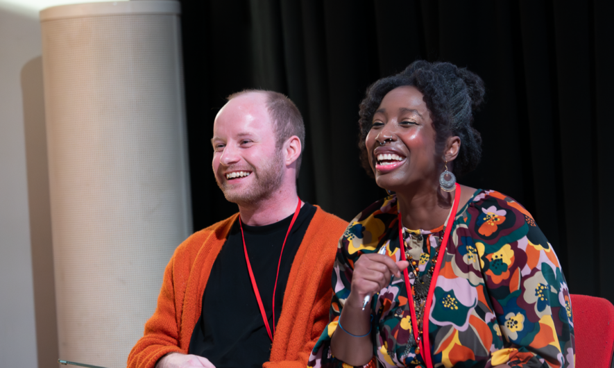 Two people sit and smile towards someone off camera.