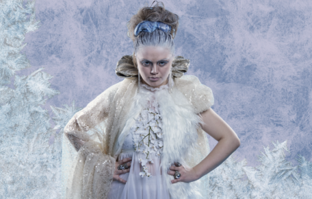 The Snow Queen stand with her hands on her hips in front of an icey background.