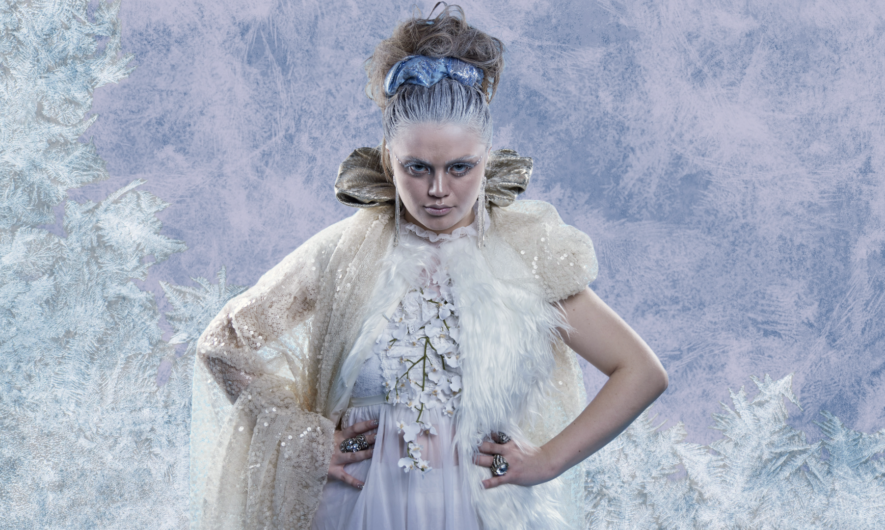 The Snow Queen stand with her hands on her hips in front of an icey background.