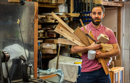 A man stands carrying planks of wood and a cup of tea, he has a biscuit in his mouth.