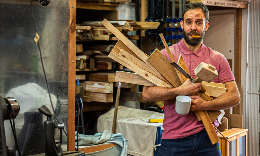 A man stands carrying planks of wood and a cup of tea, he has a biscuit in his mouth.