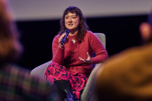 A person on stage sits and speaks into a microphone.