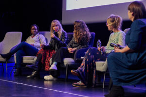 A group of people sit on a stage looking towards one speaker in the middle.
