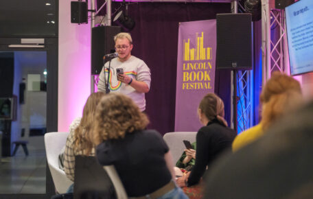 A performer stands on the Cafe stage reading from their phone.