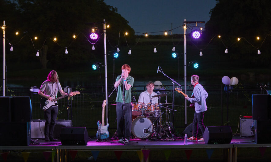 Music students perform on an outdoor stage on an evening with pink led lights behind.