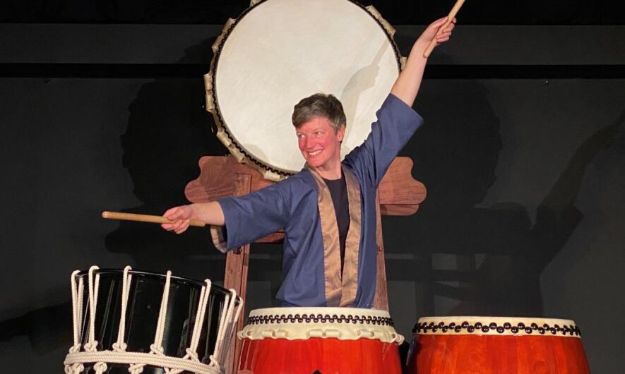 A person stands behind three large drums with their arms extended to hit two.
