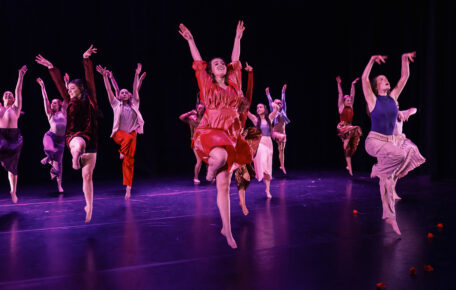 Dancers on stage jumping in the air with their arms extended into the air with their right leg bent.