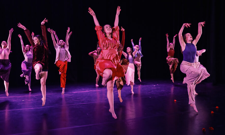 Dancers on stage jumping in the air with their arms extended into the air with their right leg bent.