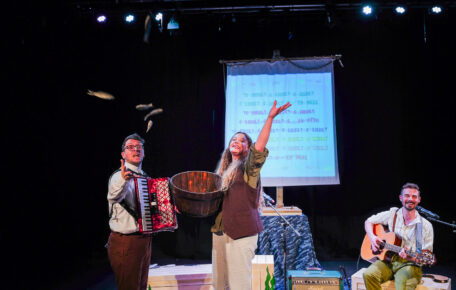 Innovate Artists Boondog Balladeers on stage during their first showing of 'Captain Godfrey and the Last Shanty' Two performers hold instruments, one with an accordion and another sat down with a guitar. Middle performer holds a wooden bucket and is throwing small fake fish towards the audience.