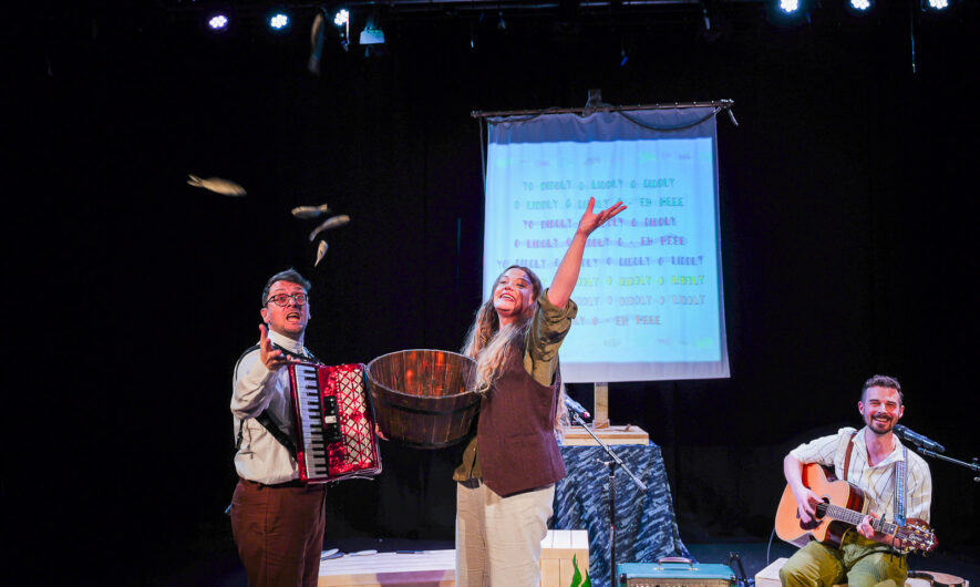 Innovate Artists Boondog Balladeers on stage during their first showing of 'Captain Godfrey and the Last Shanty' Two performers hold instruments, one with an accordion and another sat down with a guitar. Middle performer holds a wooden bucket and is throwing small fake fish towards the audience.