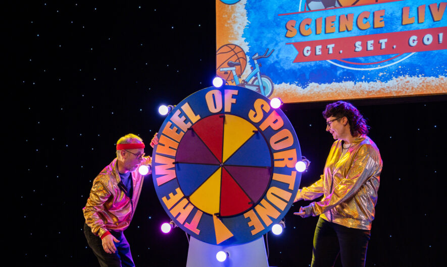 Two cast members of Science Live on stage - stood next to a spinning wheel on which is written wheel of sportune