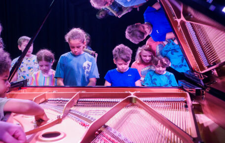 Children playing on a grand piano. The picture is taken from the back of the piano, with the lid open. There is a reflection of the children in the lid.