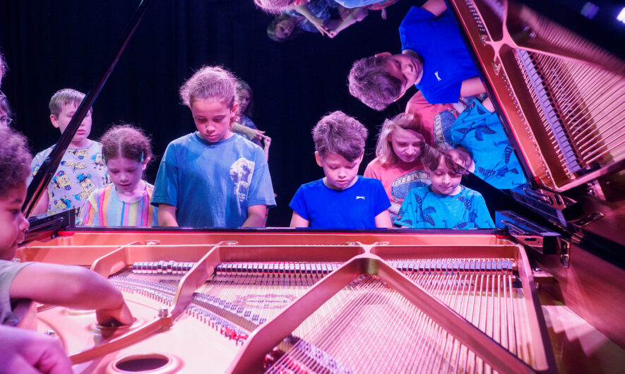 Children playing on a grand piano. The picture is taken from the back of the piano, with the lid open. There is a reflection of the children in the lid.
