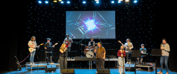 ten students are stood on stage with different instruments. Guitars, a clarinet, drum set and three singers at the front. The background is lit up with small LED light as if they were stars.