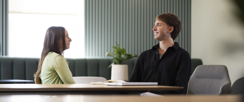 Routes In: Tom Hall is sat with Katy Gaulton in a meeting. They both have a smiling expression. Tom on the right wears a black shirt and has short brown hair. Katy wears a green cardigan and has long brown hair.
