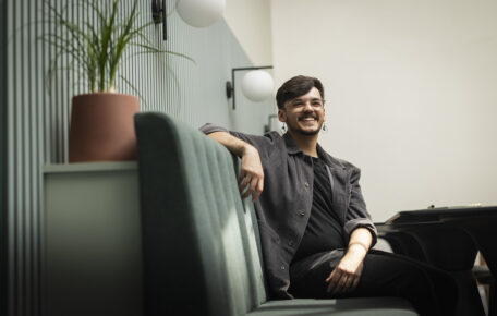 Zendon Page is seated on a blue sofa bench in the Barbican creative hub. His arm is leaning against the backrest and he has a bright smiling expression. He wears a grey overshirt with a black tshirt and trousers. On the right behind the backrest is a small plant in a terracotta plant pot.