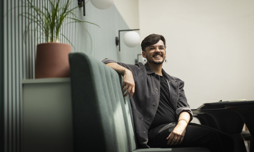 Routes In: Zendon Page is seated on a blue sofa bench in the Barbican creative hub. His arm is leaning against the backrest and he has a bright smiling expression. He wears a grey overshirt with a black tshirt and trousers. On the right behind the backrest is a small plant in a terracotta plant pot.