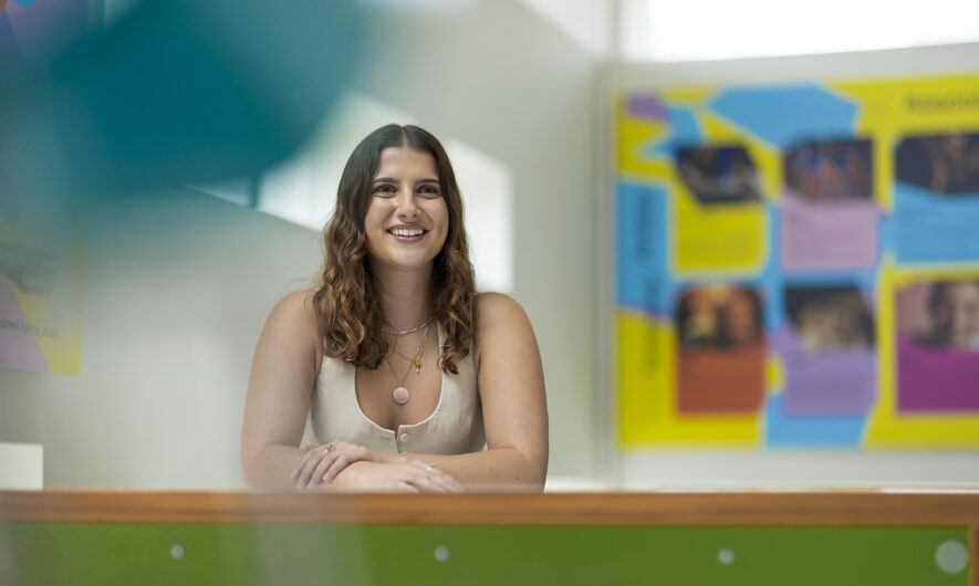 Routes In: Amber Jesson is seated in the LAC cafe. Her arms are folded on the table and she has a smiling expression. She wears a beige waistcoat with golden necklaces and a shell pendant. She has mid length brown wavy hair. In the background is the colourful artist boards and bright light coming through the window.
