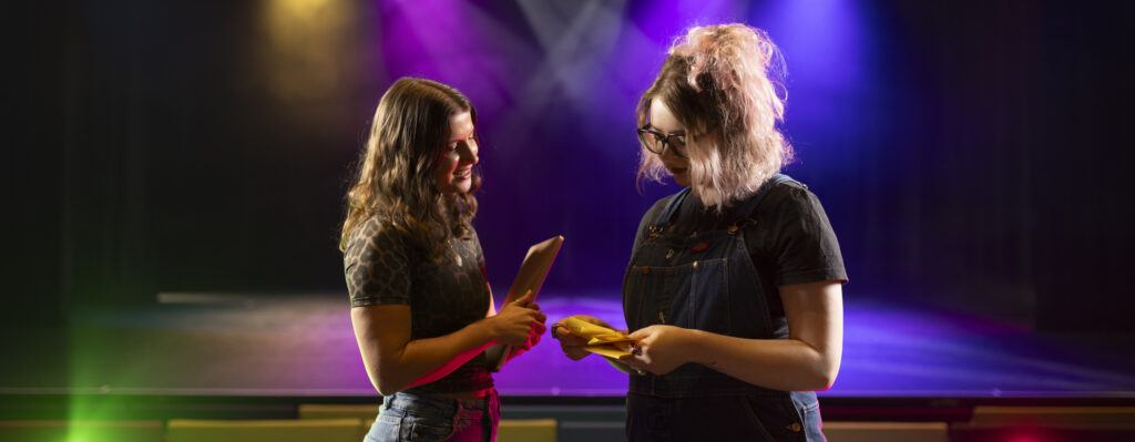 Routes In colleague Amber Jesson is stood with artist Rosie Hollingworth in front of a colourful light stage in the auditorium. They look as they are having a discussion and looking over some notes.