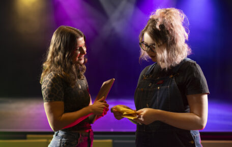 Amber Jesson is stood with artist Rosie Hollingworth in front of a colourful light stage in the auditorium. They look as they are having a discussion and looking over some notes.