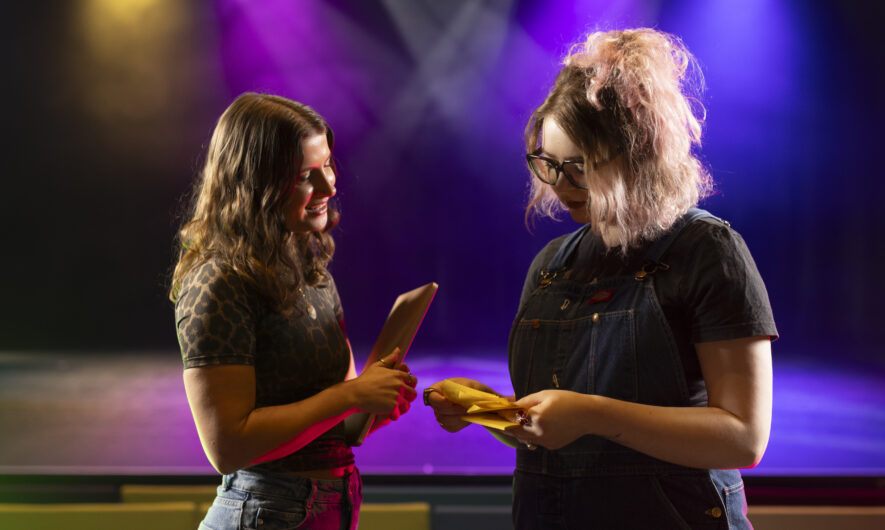 Amber Jesson is stood with artist Rosie Hollingworth in front of a colourful light stage in the auditorium. They look as they are having a discussion and looking over some notes.