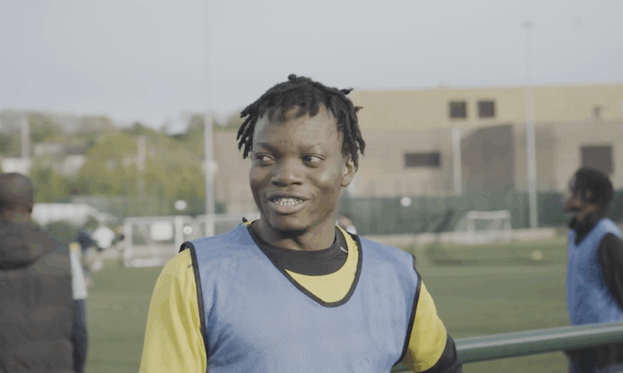 Screen shot from the Kicking Off documentary shows a young player with dark hair smiling. He wears a bright yellow tricot and blue vest.
