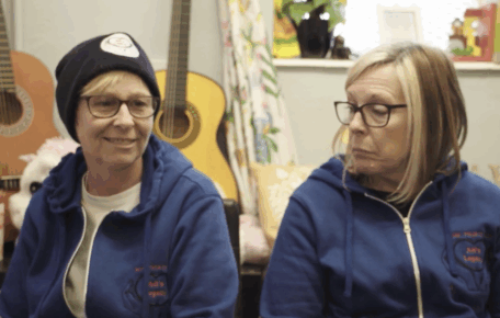 screenshot from Legacy documentary - shows two women from Adi's Legacy charity they both wear glasses and navy jumpers embroidered with the logo. On the left she has a short blond hair and wears a black beanie and on the right she had a blond bob haircut.
