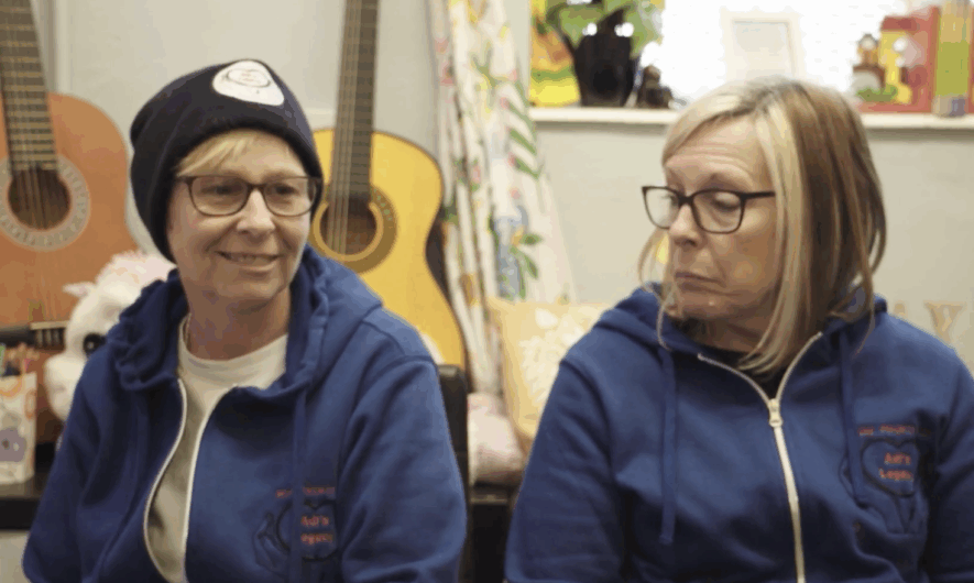 screenshot from Legacy documentary - shows two women from Adi's Legacy charity they both wear glasses and navy jumpers embroidered with the logo. On the left she has a short blond hair and wears a black beanie and on the right she had a blond bob haircut.