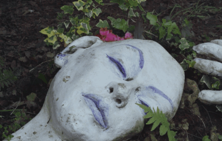 Screenshot from short film Still Dawber - shows clay sculpture in the garden of a sleeping woman enveloped by the plants. Her skin is a bright white and her features are painted blue.
