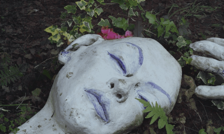 Screenshot from short film Still Dawber - shows clay sculpture in the garden of a sleeping woman enveloped by the plants. Her skin is a bright white and her features are painted blue.